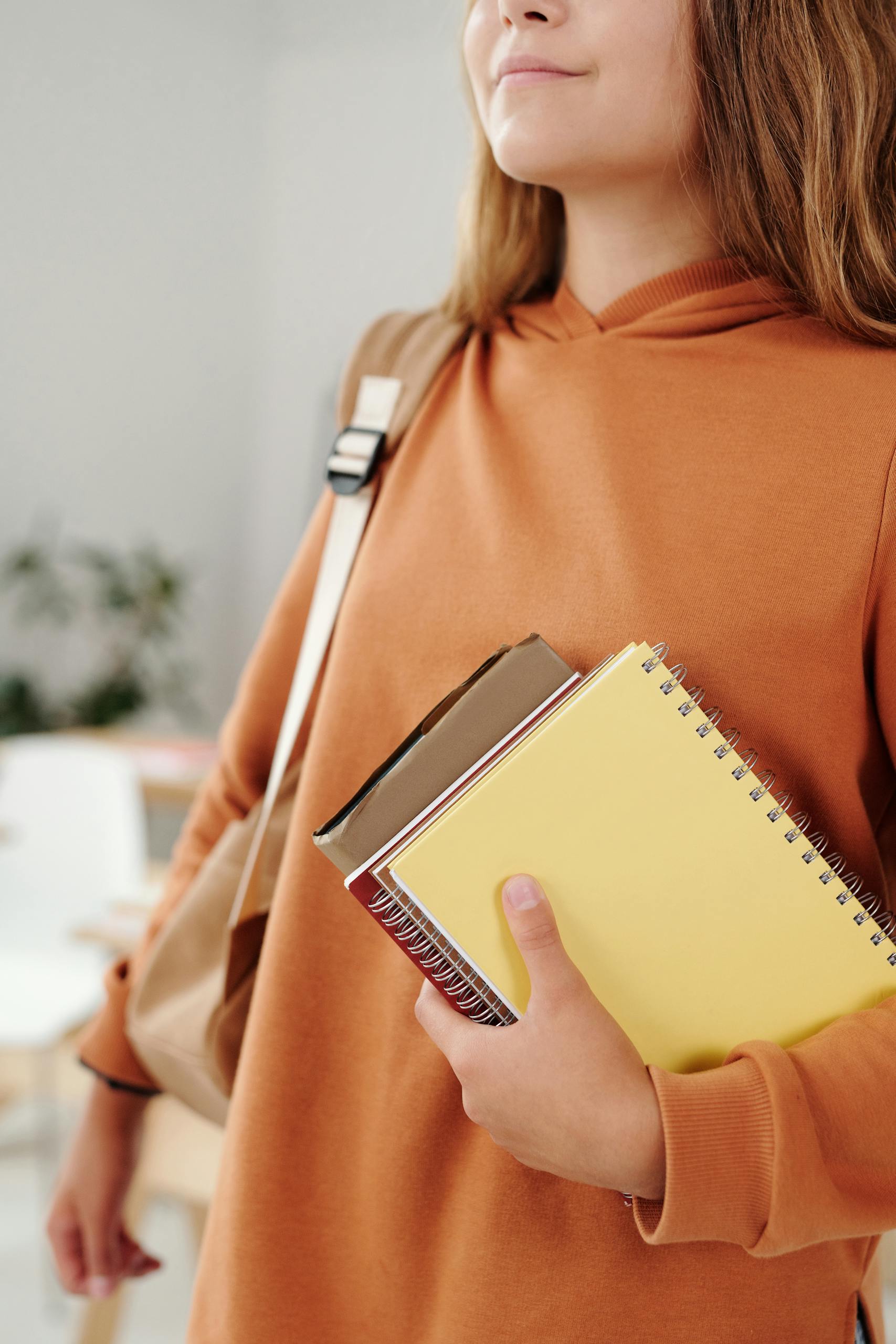 Schoolgirl in orange hoodie holding notebooks indoors, embodying education and back-to-school vibes.